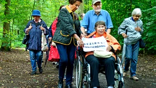 Foto: Eine Spaziergangsgruppe im Wald, im Bildvordergrund eine Dame im Rollstuhl, geschoben von ihrem Mann, begleitet zu ihrer Rechten von einer weiteren Dame, mehr Spaziergehende im Hintergrund