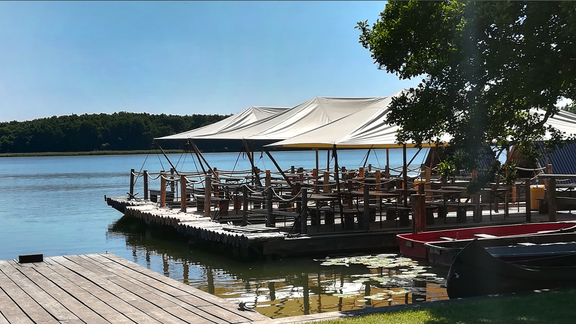 Foto: Ein Groß-Floß an seiner Anlegestelle am Seeufer, im Hintergrund der See und Wald unter blauem Himmel