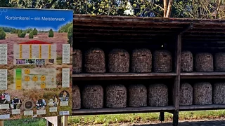 Foto: Eine doppelte Reihe an Imkerkörben, im Vordergrund eine Infotafel zum Thema