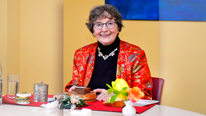 Foto: Märchenerzählerin Heike Grützmacher, eine lächelnde, ältere Dame mit bunt besticktem, roten Jacket an einem Tisch sitzend